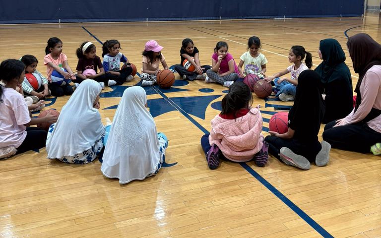 Young girls sitting in a circle with basketballs while their coaches talk with them.