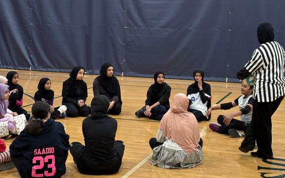 Girls sitting in a circle on a gym floor while a referee balances a volleyball on a player's head.